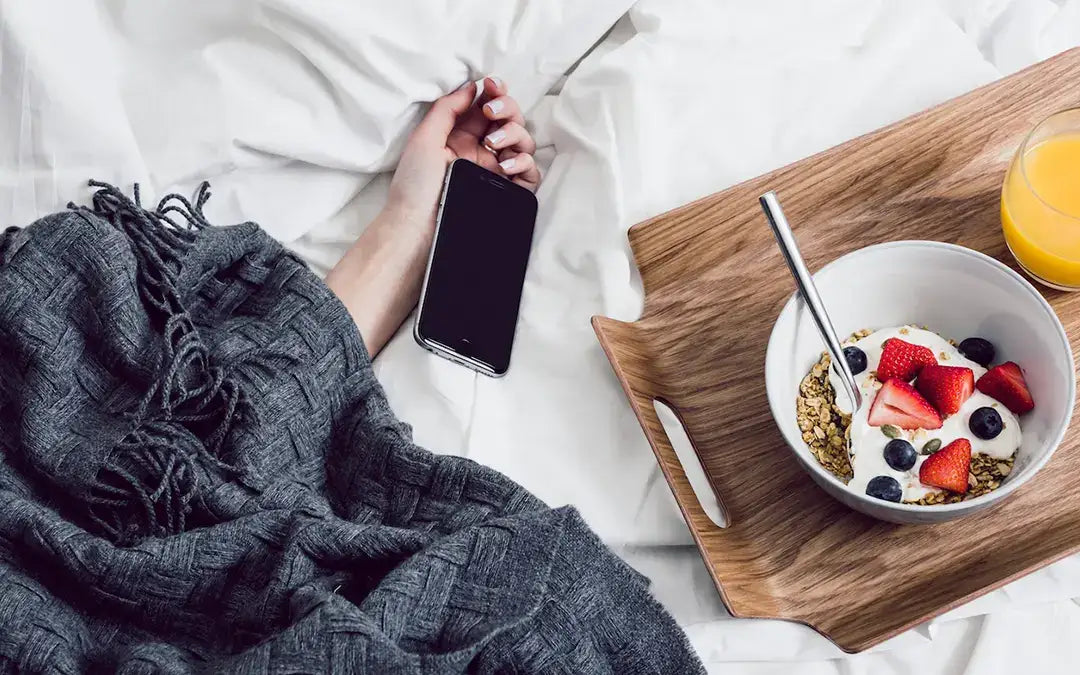 hand of sleeping woman under blanket with cell phone and tray holding bowl of yogurt with fruit and granola