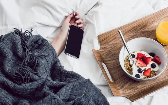 hand of sleeping woman under blanket with cell phone and tray holding bowl of yogurt with fruit and granola