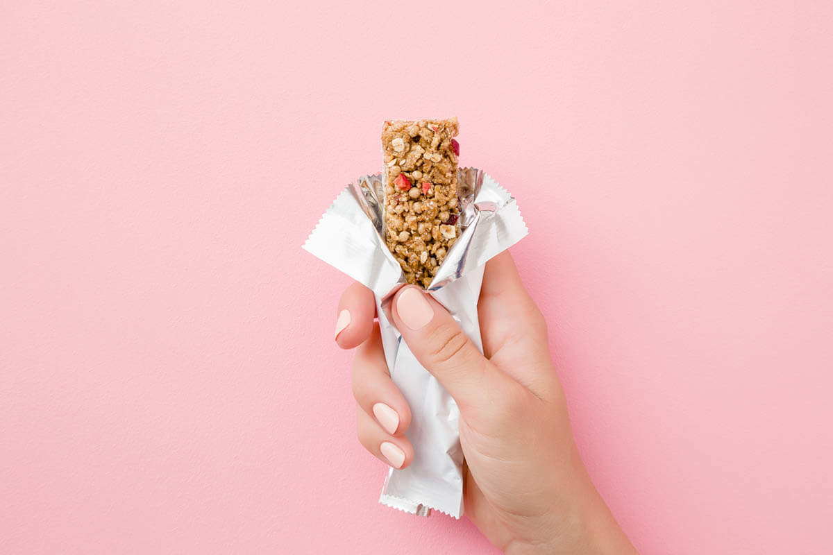 Young woman hand holding packaged cereal bar on pastel pink table. Opened white pack. Closeup. Top view.