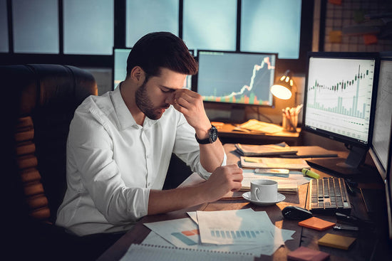 stressed young man sitting at a desk with computer screens and papers