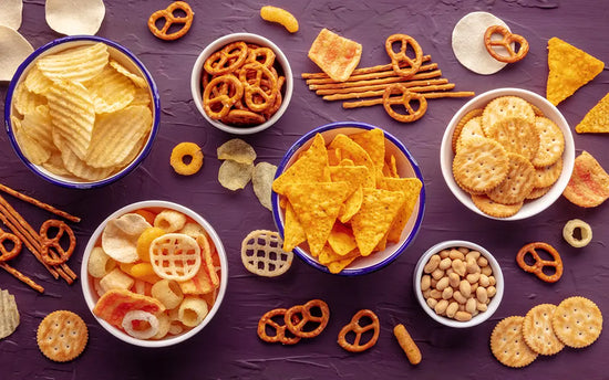 bowls of ultra processed foods like chips, snack mixes, and pretzels on a dark background