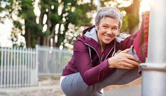 Smiling retired woman listening to music while stretching legs outdoors. Senior woman enjoying daily routine warming up before running at morning. Sporty lady doing leg stretches before workout and looking at camera.