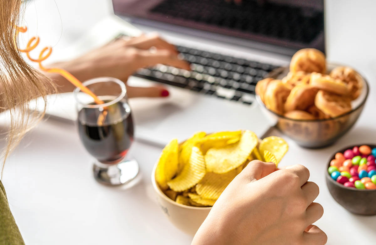 Girl works at a computer and eats unhealthy, processed food while drinking wine, all of which weaken the immune system