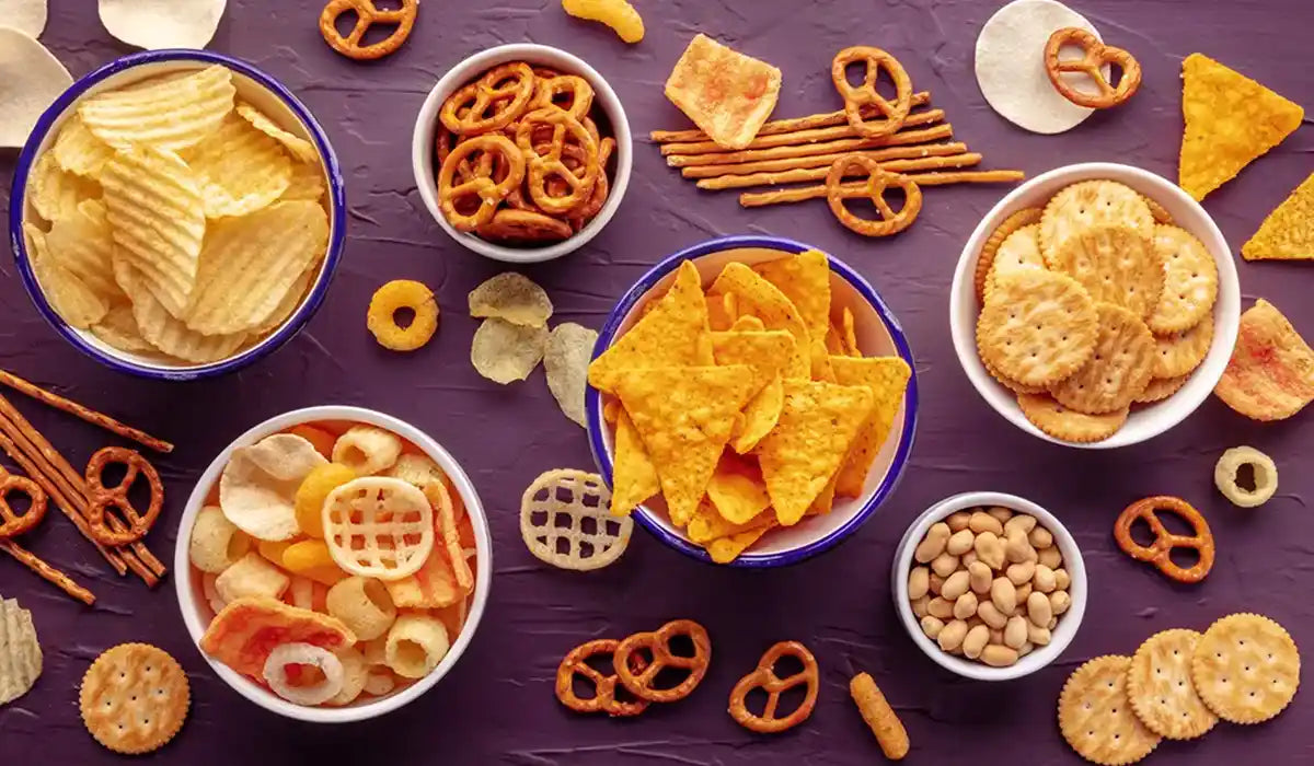 small bowls of processed chips, crackers, and pretzels on a dark surface